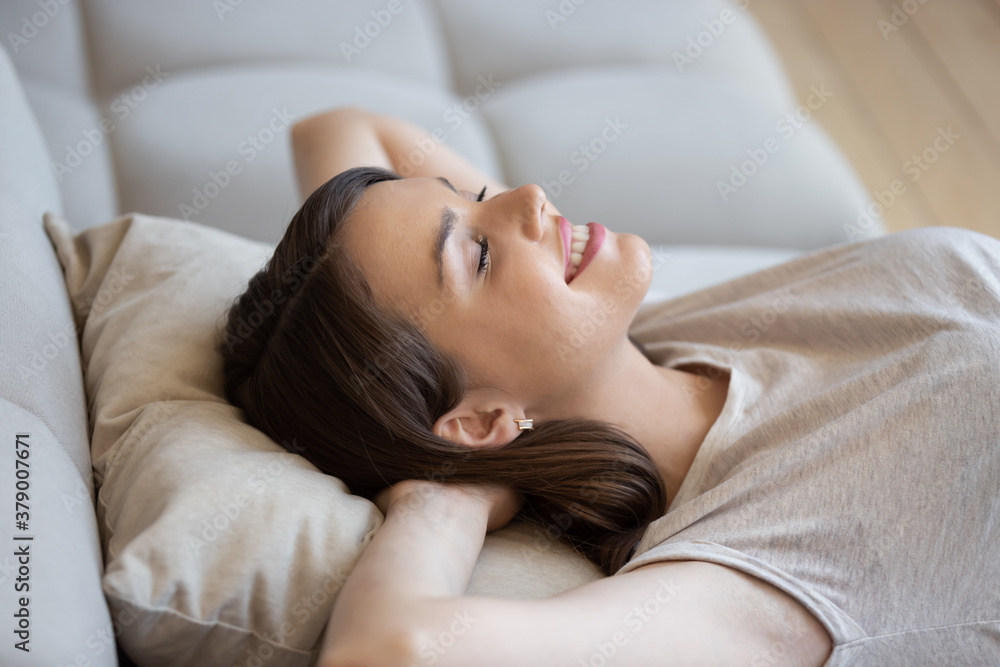 Happy relaxed woman lying on couch with eyes closed and enjoying peace and quiet of her own soundproof house. Good-looking young lady lounging on sofa cushion and smiling feeling content and fulfilled - obrazy, fototapety, plakaty 