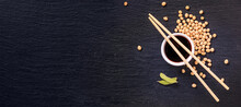 Soy sauce in a bowl with bamboo chopsticks and soya beans scattered on the black surface of the slate stone, banner, top view, closeup with space for text