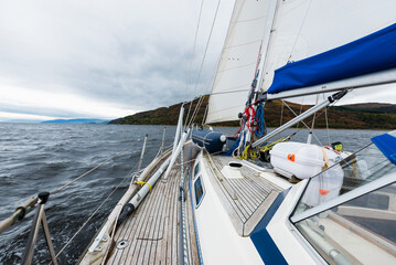 Wall Mural - Sloop rigged modern yacht with wooden teak deck sailing near the rocky shores of Tarbert after the rain. Scotland, UK. Close-up view from the deck to the bow, mast and sails. Dramatic storm sky