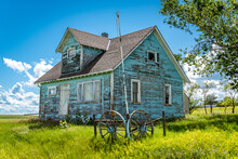 Old, Abandoned Blue Prairie Farmhouse With Trees, Grass, Blue Sky And A Wagon Wheel In Kayville, Saskatchewan, Canada