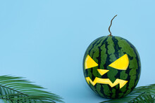 Glowing Carved Halloween Watermelon On Blue Background With Palm Leaves. Green Jack O Lantern With Copy Space.