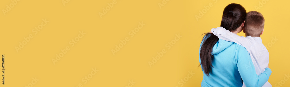 Brunette boy and mother sitting together, hugs, love, family. Isolated on yellow background, copy space template, banner. - obrazy, fototapety, plakaty 