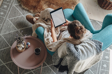 woman sitting in cozy armchair, with warm blanket, using tablet