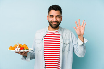 Wall Mural - Young latin man holding a waffle isolated smiling cheerful showing number five with fingers.