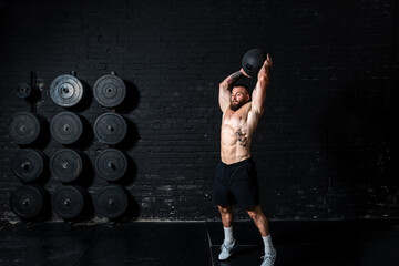 Young sweaty strong muscular fit man with big muscles doing ball throwing on the floor as hardcore cross workout training in the gym