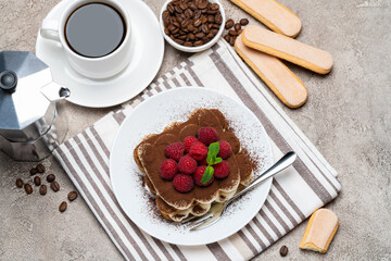 portion of Classic tiramisu dessert with raspberries, cup of espresso, coffee maker on grey concrete background