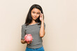 © Asier - Young asian woman holding a piggy bank pointing temple with finger, thinking, focused on a task.