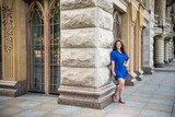 Fototapeta Na drzwi - Beautiful young smiling woman walking in city park near famous old architecture building at summer day wearing dress and looking to the camera