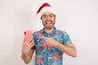 © Roquillo - Portrait of Young caucasian man wearing hawaiian shirt and Santa hat over isolated white background holding in hands showing new cell,