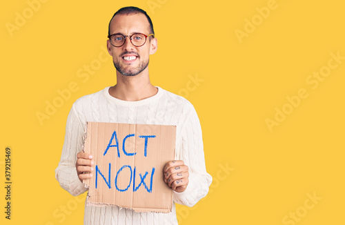 Fotomural Young handsome man holding act now banner looking positive and happy standing an