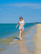 © Nekrasov - A young girl runs along the sandy sea beach along the surf. Splashes of sea water. The girl is wearing a white swimsuit and sunglasses. Freedom and carelessness. Outdoor fitness. Sunny day. Copy space