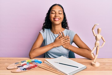 Young african american girl artist sitting at studio table smiling with hands on chest, eyes closed with grateful gesture on face. health concept.