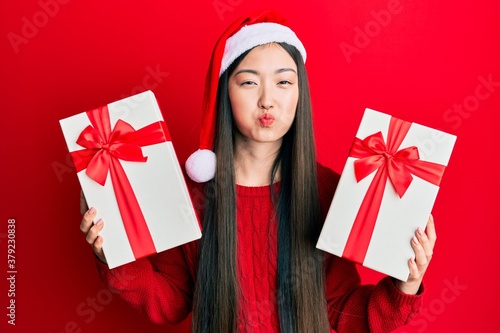 Fotomural Young chinese woman wearing christmas hat and holding gifts puffing cheeks with funny face