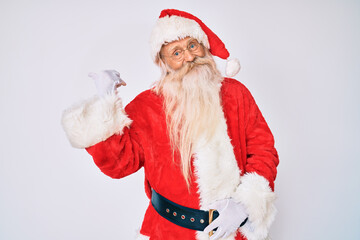 Old senior man with grey hair and long beard wearing santa claus costume with suspenders smiling with happy face looking and pointing to the side with thumb up.