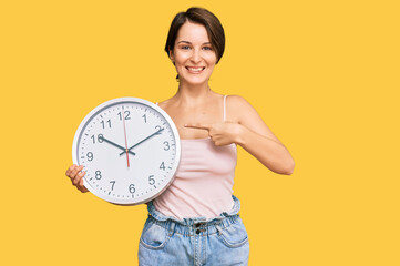 Young brunette woman with short hair holding big clock smiling happy pointing with hand and finger