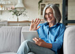 © insta_photos - Happy 60s older mature middle aged adult woman waving hand holding digital tablet computer video conference calling by social distance virtual family online chat meeting sitting on couch at home.