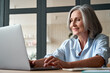© insta_photos - Smiling stylish mature middle aged business woman using laptop computer sitting at workplace desk. Happy senior older lady, 60s grey-haired businesswoman working typing on pc at home or from office.