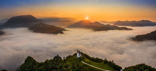 Wall Mural - Sebrelje, Slovenia - Aerial panoramic drone view of the beautiful hilltop church of St.Ivan (Sv. Ivan Cerkev) at sunrise with huge morning fog bellow the valley and Julian Alps at background