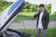 Young Asian man worries using mobile phone calling for assistance with his car broken down by the roadside. Open the hood to see about the engine and explain the mechanic the problem with automobile.