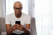 © DragonImages - Smiling young Black man in glasses sitting on sofa and checking news on social media