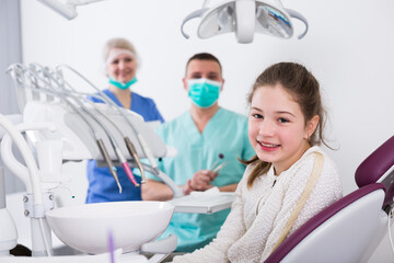 Happy teen girl sitting in dental chair after teeth cure with two professional dentists behind