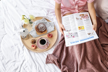 Wall Mural - Young woman reading newspaper while having tasty breakfast in bed