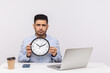 © khosrork - Time management. Serious displeased man employee sitting in office workplace with laptop, holding big clock