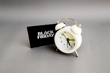 white alarm clock and an inscription on a black square - black Friday on a gray background. The topic of sales.