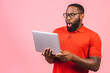 Young surprised shocked amazed african man standing and using laptop computer isolated over pink background.