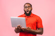 © denis_vermenko - Young surprised shocked amazed african man standing and using laptop computer isolated over pink background.