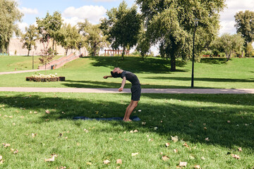 Young athletic man in sportswear doing yoga in the park. Practice asana outdoors. People exercising on green grass with yoga mat. Strong mature caucasian man in dsun salutations pose