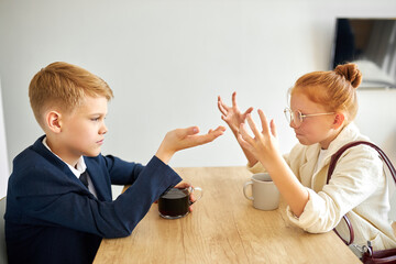 cute kids quarelling at home sitting at table, discuss their relationships, in formal wear. redhead girl emotionally say something