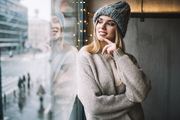 Canvas Print - Contemplative female teenager dressed in stylish winter hat and knitted sweater looking at window and dreaming, happy Caucasian hipster girl 20 years old enjoying recreation and youth lifestyle