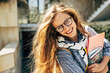 © iuricazac - Outdoor portrait of a happy young student woman wearing transparent eyeglasses looking to the camera, standing next to the college campus and carrying lots of books and folders on a sunny day.