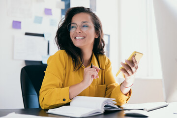 Pretty cheerful businesswoman looking away sitting at desk and holding phone in hand. Positive thinking person concept. Notes and plans on background. Female office worker of freelancer.