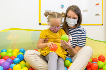 Toddler girl in child occupational therapy session doing playful exercises with her therapist during Covid - 19 pandemic, both wearing protective face masks.