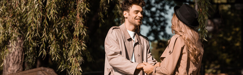 panoramic crop of man in trench coat looking at woman in hat while holding hands