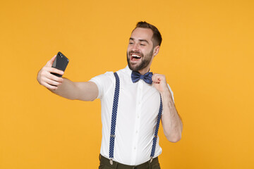 Wall Mural - Funny cheerful laughing young bearded man 20s wearing white shirt bow-tie suspender posing standing doing selfie shot on mobile phone isolated on bright yellow color wall background studio portrait.