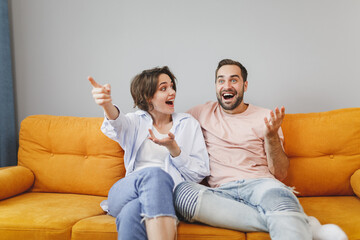 Canvas Print - Shocked excited young couple two friends man woman 20s in casual clothes sitting on couch hugging spreading hands pointing index finger aside resting relaxing spending time in living room at home.