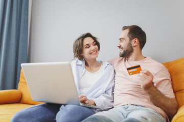 Wall Mural - Smiling cheerful pretty young couple two friends man woman 20s in casual clothes sitting on couch using laptop pc computer hold credit bank card resting relaxing spending time in living room at home.