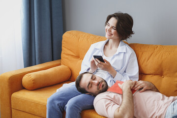 Poster - Funny smiling young couple two friends man woman 20s wearing casual clothes sitting lying on couch using mobile cell phone reading book sleeping resting relaxing spending time in living room at home.