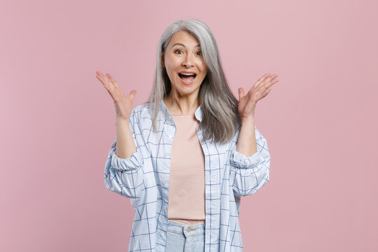 Shocked amazed excited surprised gray-haired asian woman in basic white checkered shirt spreading hands keeping mouth open looking camera isolated on pastel pink colour background, studio portrait.