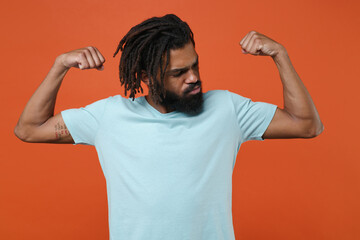 Wall Mural - Strong young african american man guy wearing blue casual t-shirt posing isolated on bright orange wall background studio portrait. People sincere emotions lifestyle concept. Showing biceps, muscles.