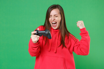 Wall Mural - Screaming overjoyed young brunette woman 20s wearing bright red casual streetwear hoodie posing playing game with joystick doing winner gesture isolated on green color wall background studio portrait.