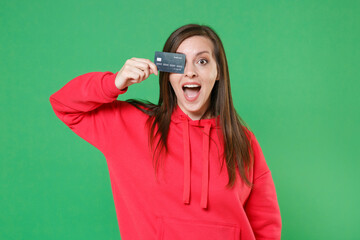 Wall Mural - Surprised cheerful young brunette woman 20s wearing bright red casual streetwear hoodie posing covering eye with credit bank card keeping mouth open isolated on green color background studio portrait.