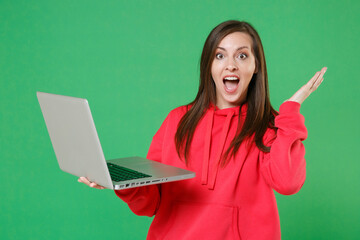 Wall Mural - Surprised young brunette woman 20s in bright red casual streetwear hoodie posing working on laptop pc computer spreading hands keeping mouth open isolated on green color background studio portrait.