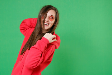 Wall Mural - Side view of smiling cheerful laughing beautiful young brunette woman 20s in bright red casual streetwear hoodie eyeglasses posing looking aside isolated on green color background studio portrait.