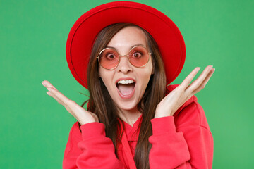 Wall Mural - Excited surprised young brunette woman 20s in red casual streetwear hoodie eyeglasses hat posing keeping mouth open spreading hands looking camera isolated on green color background studio portrait.