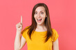 Excited shocked young brunette woman 20s wearing yellow casual t-shirt posing standing holding index finger up with great new idea looking camera isolated on pink color background studio portrait.