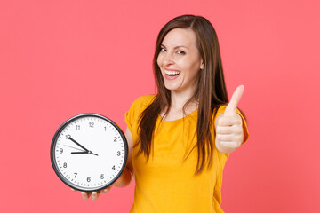 Wall Mural - Smiling cheerful laughing young brunette woman 20s in yellow casual t-shirt posing standing holding in hands clock showing thumb up looking camera isolated on pink color background studio portrait.